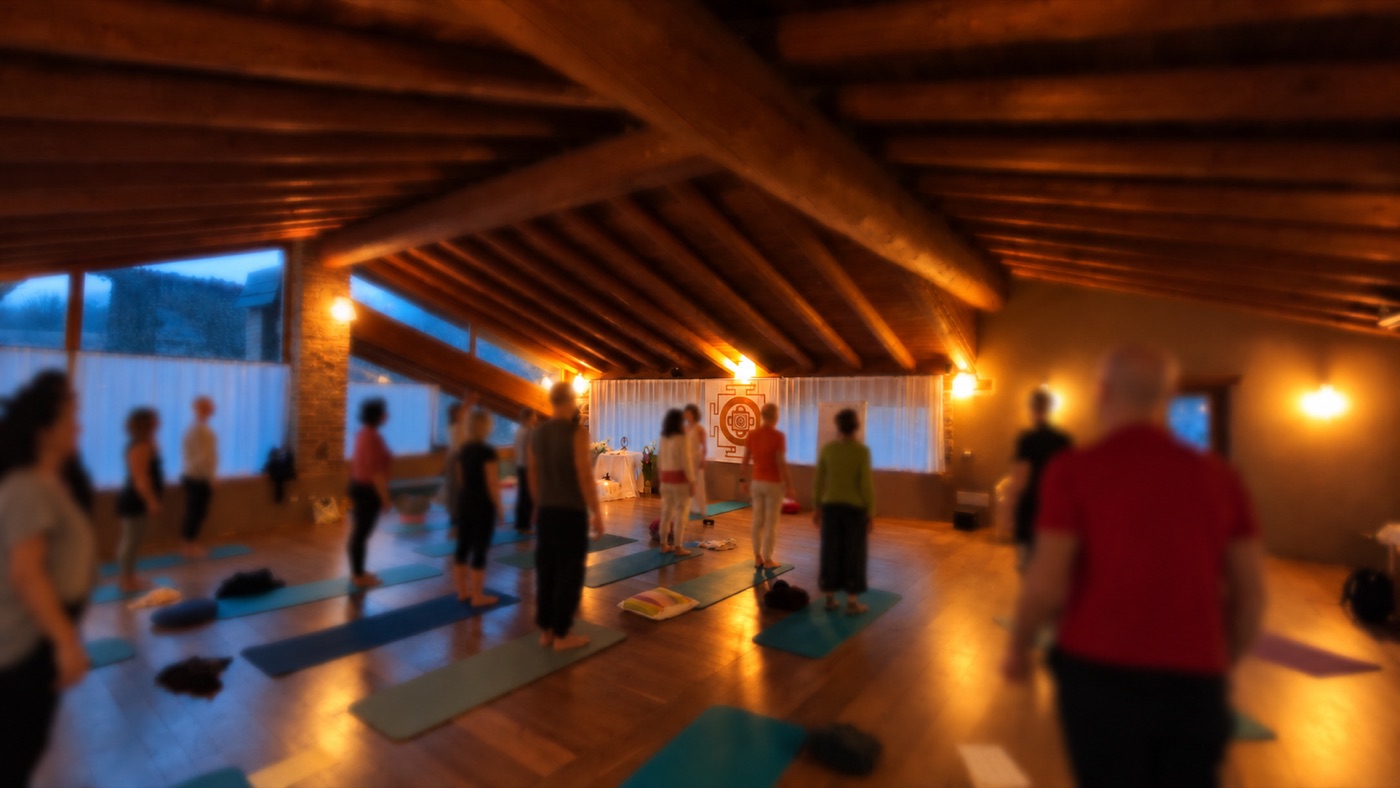 Un gruppo di partecipanti in piedi sui tappetini durante una pratica di Tantra Yoga, in una sala con travi in legno e un mandala appeso alla parete