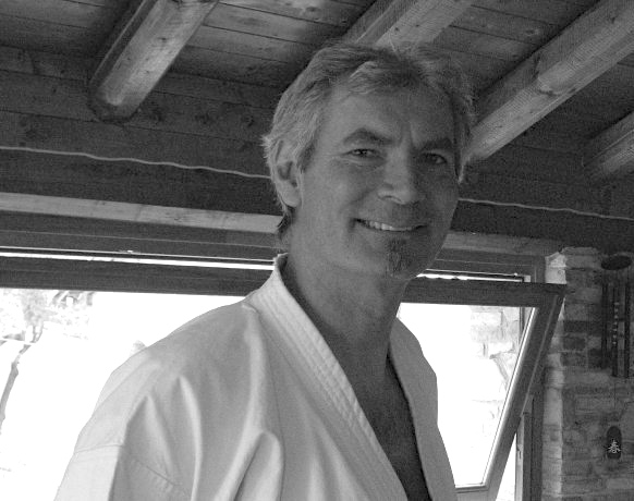 Carlo Pedrazzini, sorridente, con il karategi bianco, sotto un porticato con travi di legno (foto in bianco e nero)